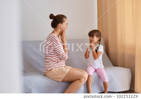Side view of woman speech pathologist having lesson with little girl, language therapist practicing sounds pronunciation with kid, people sitting on comfortable sofa near window. 102251229