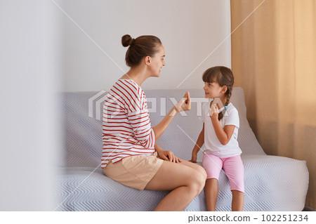 Woman wearing striped shirt and shorts sitting on sofa with little girl, articulation exercised at lesson at home, mother or teacher and female kid learn pronouncing sounds together. 102251234
