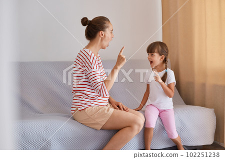 Indoor shot of hard working woman speech therapist working with little girl at home, female wearing striped shirt and shorts sitting on sofa with child. Indoor shot of hard working woman speech therapist working with little girl at home, female wearing striped shirt and shorts sitting on sofa with child. 102251238