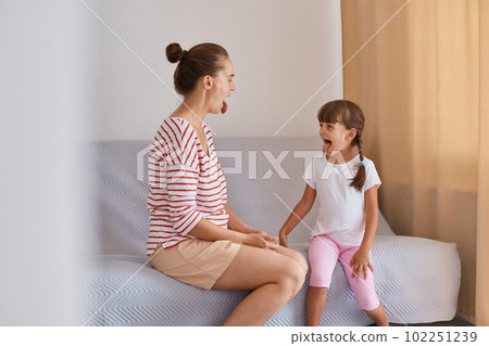 Horizontal shot of Caucasian woman wearing striped shirt and shorts sitting on sofa with little girl, doing exercises with tongue out, learning pronouncing sounds together. 102251239