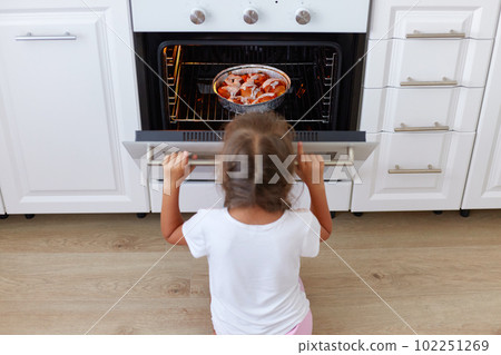 Charming girl sitting near gas stove on floor in kitchen and opening oven, curious female child waiting for cake or pie, baking in stove, homemade pastry. Charming girl sitting near gas stove on floor in kitchen and opening oven, curious female child waiting for cake or pie, baking in stove, homemade pastry. 102251269
