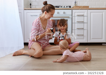 Indoor shot of charming family spending time together while baking together, woman wearing striped shirt sitting on floor in kitchen with her little daughters. Indoor shot of charming family spending time together while baking together, woman wearing striped shirt sitting on floor in kitchen with her little daughters. 102251328