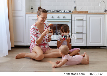 Portrait of attractive female with hair ban wearing striped casual shirt sitting on floor in kitchen with cup in hands, spending time with her little daughters. Portrait of attractive female with hair ban wearing striped casual shirt sitting on floor in kitchen with cup in hands, spending time with her little daughters. 102251329