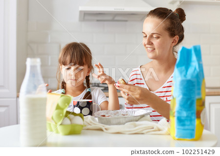 Portrait of optimistic happy woman sitting at table with her daughter and eating tasty cakes, posing in kitchen, expressing positive emotions and happiness. 102251429