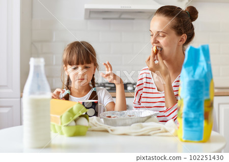 Image of happy woman biting homemade cake, sitting at table with her charming daughter and eating tasty pie, posing in kitchen, tasting fresh baked pastries. Image of happy woman biting homemade cake, sitting at table with her charming daughter and eating tasty pie, posing in kitchen, tasting fresh baked pastries. 102251430