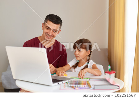 Smiling father and a little daughter doing homework at home, sitting at table in room, looking at laptop screen with smiles, having positive emotions, online education. 102251567