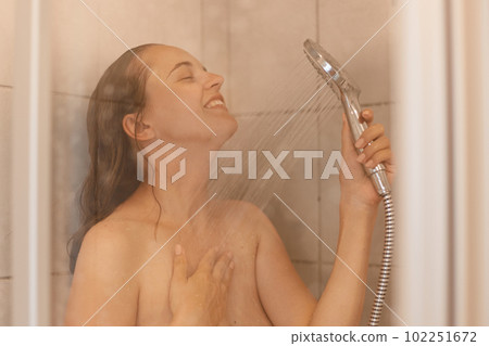 Indoor shot of beautiful young adult woman with naked body taking shower, standing under water with satisfied facial expression, enjoying relaxation in bathroom. 102251672
