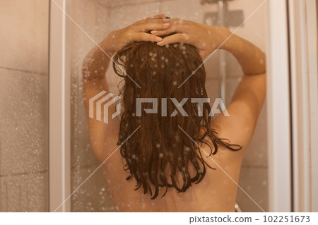 Back side photo of dark haired woman with naked body takes shower behind glass door in bathroom, enjoys relaxation in bathroom, has healthy pure skin. Back side photo of dark haired woman with naked body takes shower behind glass door in bathroom, enjoys relaxation in bathroom, has healthy pure skin. 102251673