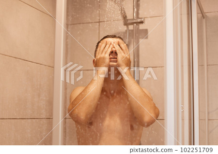 Portrait of young brunette relaxed man taking shower, standing under falling water drops, washing body and face in bathroom at home, bodycare and everyday hygiene routine. 102251709