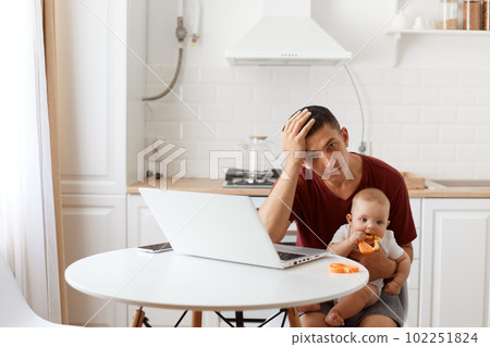 Tired exhausted man handsome freelancer male wearing burgundy r shirt, posing in white kitchen, sitting in front of laptop with baby in hands, having headache. Tired exhausted man handsome freelancer male wearing burgundy r shirt, posing in white kitchen, sitting in front of laptop with baby in hands, having headache. 102251824
