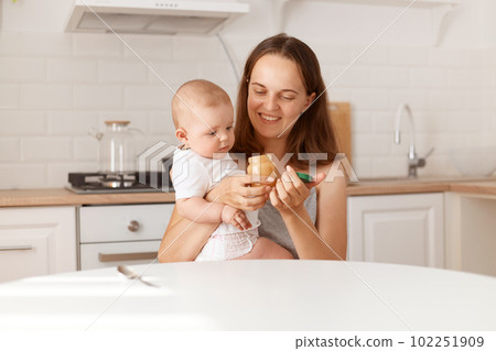 Happy dark haired female mother opens jar with fruit or vegetable puree with her daughter, expressing positive emotions, sitting at table in white kitchen. Happy dark haired female mother opens jar with fruit or vegetable puree with her daughter, expressing positive emotions, sitting at table in white kitchen. 102251909