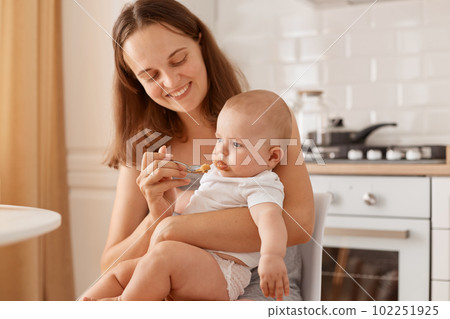 Dark haired female feeding her little daughter with fruit or vegetable puree, looking at baby with charming smile, holding spoon in hand, looking at toddler girl. Dark haired female feeding her little daughter with fruit or vegetable puree, looking at baby with charming smile, holding spoon in hand, looking at toddler girl. 102251925