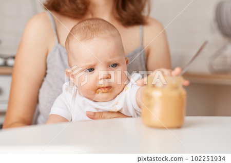 Closeup portrait of cute adorable kid wearing white t shirt, touching jar with food with interest, faceless mother feeding her little infant daughter with vegetable puree. Closeup portrait of cute adorable kid wearing white t shirt, touching jar with food with interest, faceless mother feeding her little infant daughter with vegetable puree. 102251934