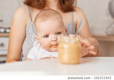 Indoor shot of cute baby in white t shirt sitting with mommy, unknown woman holding feeding infant baby with puree, posing in light kitchen at home, parenthood. 102251935
