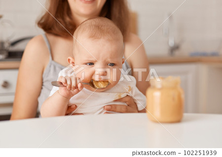 Faceless mother feeds her little infant daughter with vegetable puree, cute child trying to eat by her own, eating tasty healthy food, feeding up, indoor shot. 102251939