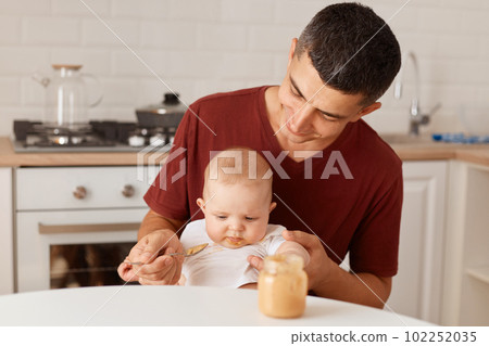Indoor shot of handsome father wearing burgundy t shirt with charming infant daughter. man feeding his baby with puree, posing in light room against kitchen set. 102252035
