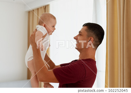 Side view of smiling happy father standing near window in light living room and holding his little daughter or son, man spending time, playing with baby at home. 102252045