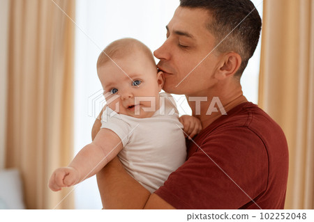 Indoor shot of young adult male wearing maroon casual style t shirt holding little cute infant girl in hands, male hugging and kissing his daughter, posing near window with beige curtains. 102252048