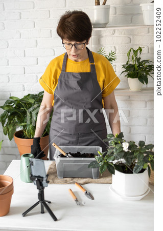 Female blogger sits in front of smartphone camera on tripod records instructional tutorial video for her blog shoots process of replanting flowers and green plants full of soil enjoys botanic hobby 102252409