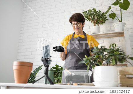Relaxing home gardening. Smiling middle aged woman in black gloves with potted plant records gardening video blog in modern house - blogging and florist vlog influencer 102252417