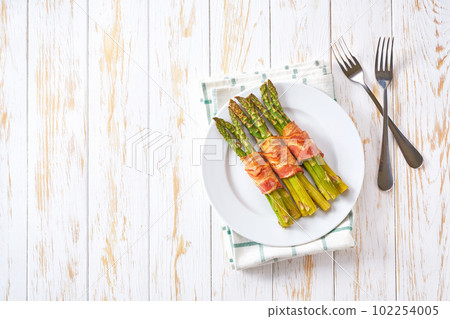 Baked bundles of green asparagus wrapped in bacon in a ceramic plate on a white wooden table, top view. Baked bundles of green asparagus wrapped in bacon in a ceramic plate on a white wooden table, top view. 102254005