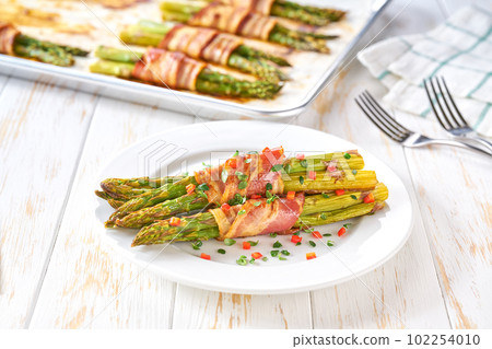 Traditional appetizer, baked  bundles of green asparagus wrapped in bacon in a ceramic plate on a white wooden table, close up. 102254010