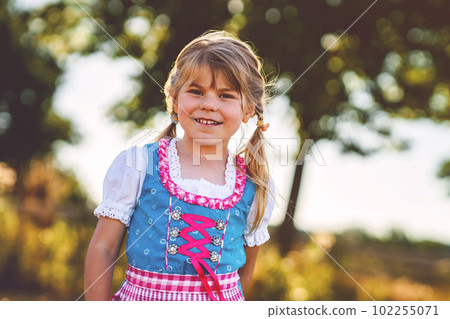 Cute little kid girl in traditional Bavarian costume in wheat field. Happy child with hay bale during Oktoberfest in Munich. Preschool girl play at hay bales during summer harvest time in Germany. 102255071