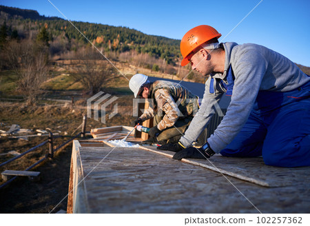 Male builder doing thermal insulation on roof of wooden frame house. Man worker spraying polyurethane foam on rooftop of future cottage. Construction and insulation concept. 102257362