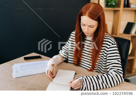High-angle view portrait of injured young woman with broken right hand wrapped in white gypsum bandage writing on paper notepad sitting at table. Concept of social security and health insurance. 102259596