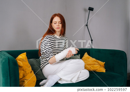 Portrait of suffering from pain injured young woman with broken right hand wrapped in white gypsum bandage sitting on couch with closed eyes. Sad redhead female with broken arm from accident at home. 102259597