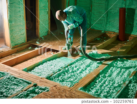 Male builder insulating wooden frame house. Man worker spraying polyurethane foam on floor inside of future cottage, using plural component gun. Construction and insulation concept. 102259761