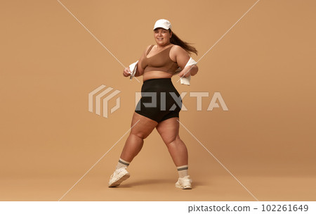 Young, positive, smiling, overweight woman posing in stylish, trendy sportswear with cap against brown studio background 102261649