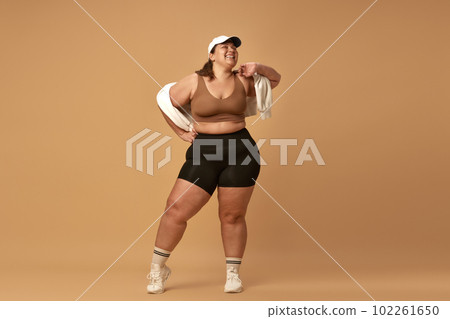Young, positive, smiling, overweight woman posing in stylish, trendy sportswear with cap against brown studio background Young, positive, smiling, overweight woman posing in stylish, trendy sportswear with cap against brown studio background 102261650