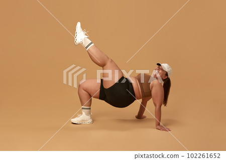 Positive, young, overweight woman training in stylish and comfortable sportswear against brown studio background. Full-body workout 102261652