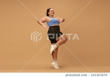 Young, positive woman with overweight figure training in sportswear against brown studio background. Cardio exercises 102261674