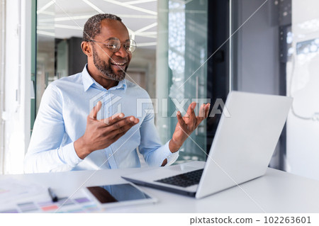 Mature african american boss working inside office with laptop remotely, businessman in shirt on online meeting with colleagues, man talking on video call using laptop while sitting in shirt. 102263601