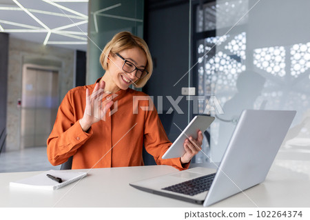 Young female student sitting in the office, campus at the table, studying and talking on a video call from the tablet. Greets, waves, smiles. Young female student sitting in the office, campus at the table, studying and talking on a video call from the tablet. Greets, waves, smiles. 102264374