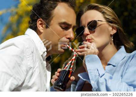 Young happy couple outdoors drinking cold drink from glass bottle with straw. Side view of man and woman drinking soft drink and smiling. 102265226