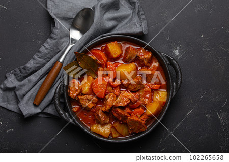 Beef meat stew with potatoes, carrot and delicious gravy in black casserole pot with bay leaves with spoon on black dark rustic concrete background from above . 102265658