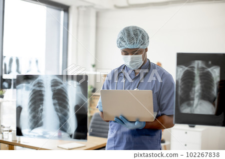 Doctor in mask and scrubs examining X-ray scans via laptop Doctor in mask and scrubs examining X-ray scans via laptop 102267838