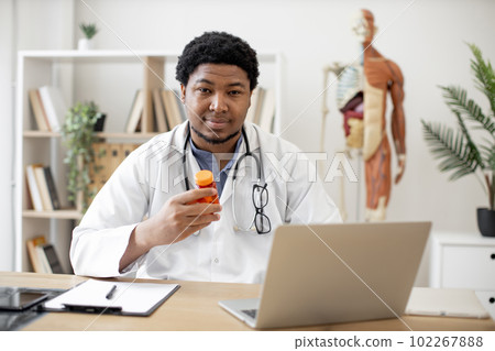 Pharmacist with pill bottle seating behind laptop in clinic Pharmacist with pill bottle seating behind laptop in clinic 102267888