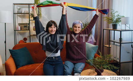 Two lesbian women family couple or girl friends holding LGBT people gay pride flag in hands at home 102267989