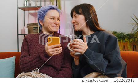 Two young cheerful lesbian friends hold smartphones, shopping on e-commerce apps at home living room Two young cheerful lesbian friends hold smartphones, shopping on e-commerce apps at home living room 102268015