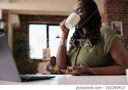 Student at home relaxing using laptop for streaming online video while drinking coffee or tea in living room. African american woman spending free time watching video content on portable computer. 102269413
