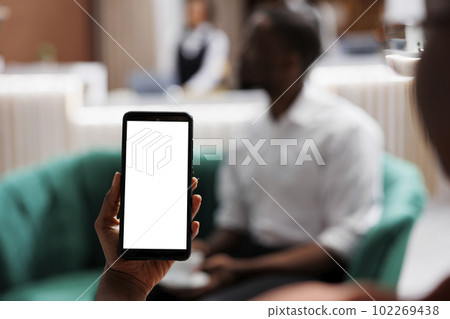 Person holding phone with blank screen, relaxing on couch in lounge area at luxury resort. Young woman guest using smartphone with white display and isolated copyspace in hotel. 102269438