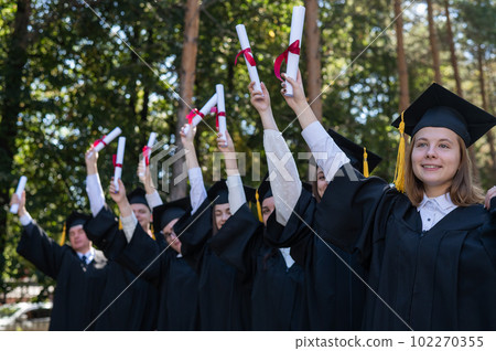 A group of graduates in robes raised their hands with diplomas outdoors. Elderly student. 102270355