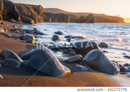 Scenic beach with black sand by low tide at sunset, Sharp mountain volcanic cliffs on Atlantic ocean coastline, Famous touristic place in Iceland 102271776