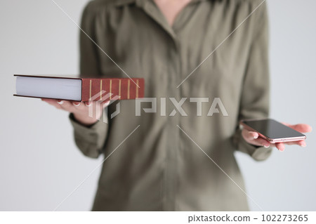 Confused woman holds book and smartphone making choice Confused woman holds book and smartphone making choice 102273265