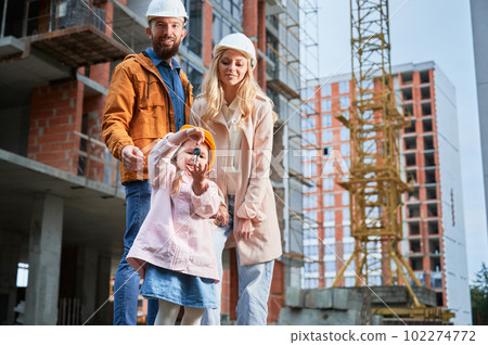 Daughter holding apartment keys and smiling while standing next to parents outside building under construction. Happy family homeowners posing on the street at construction site. 102274772