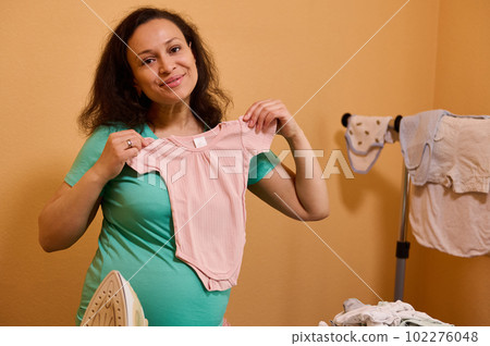 Happy expectant mother, pregnant woman smiling looking at camera, holding laundered clean ironed pink bodysuit for her newborn baby, standing by ironing board in dressing room. Household. Housekeeping 102276048